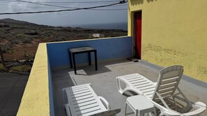 A balcony with two white plastic chairs and a small table, overlooking a hilly landscape with a view of the sea.