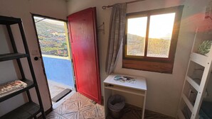 A small room with a red door, a window with curtains, a white table with a picture, a trash bin, and a view of a hillside. - 7 Calcosas (Valverde)