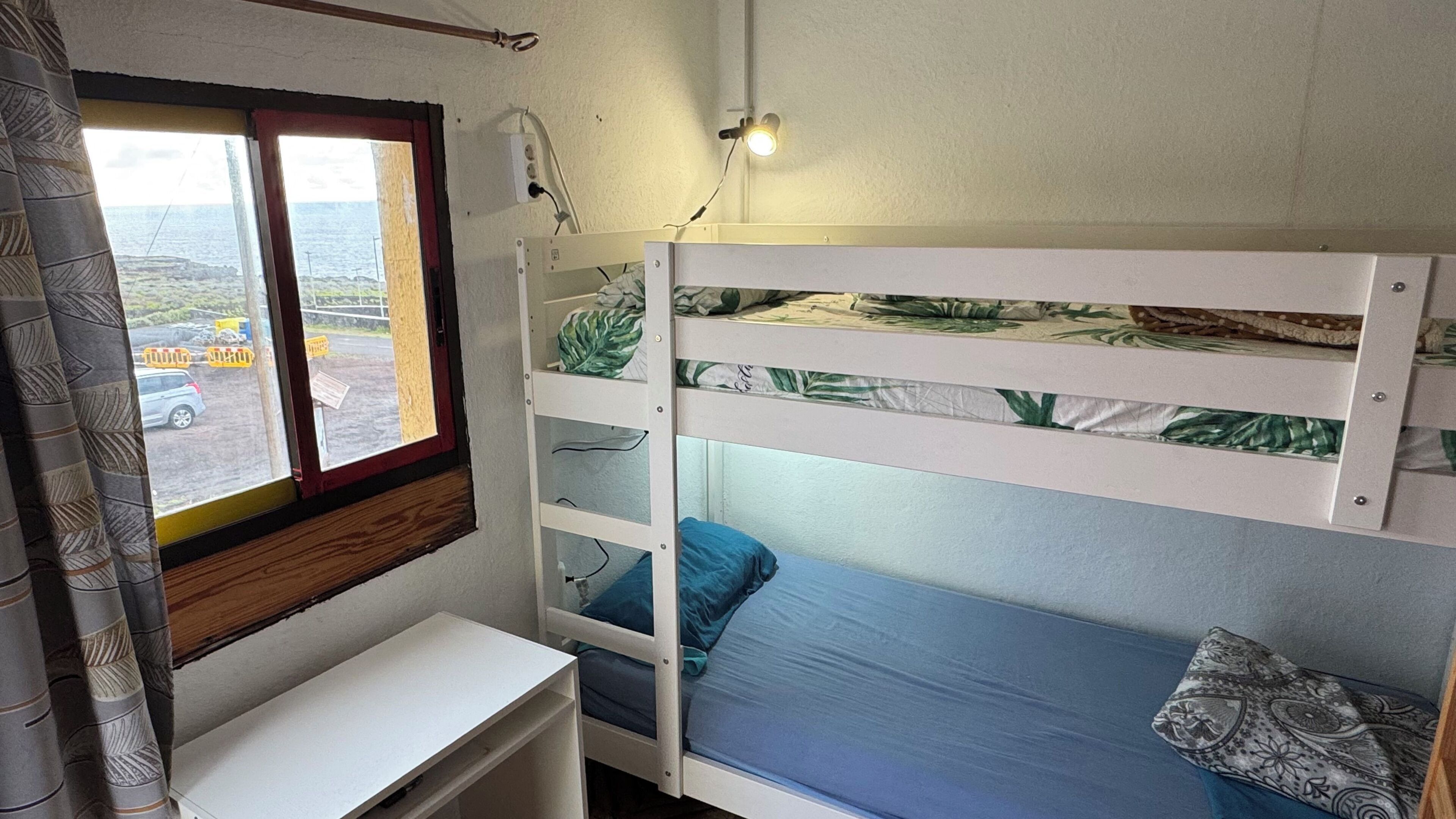 A bunk bed with a desk, a window with a view of the outside, and a wall-mounted light fixture.