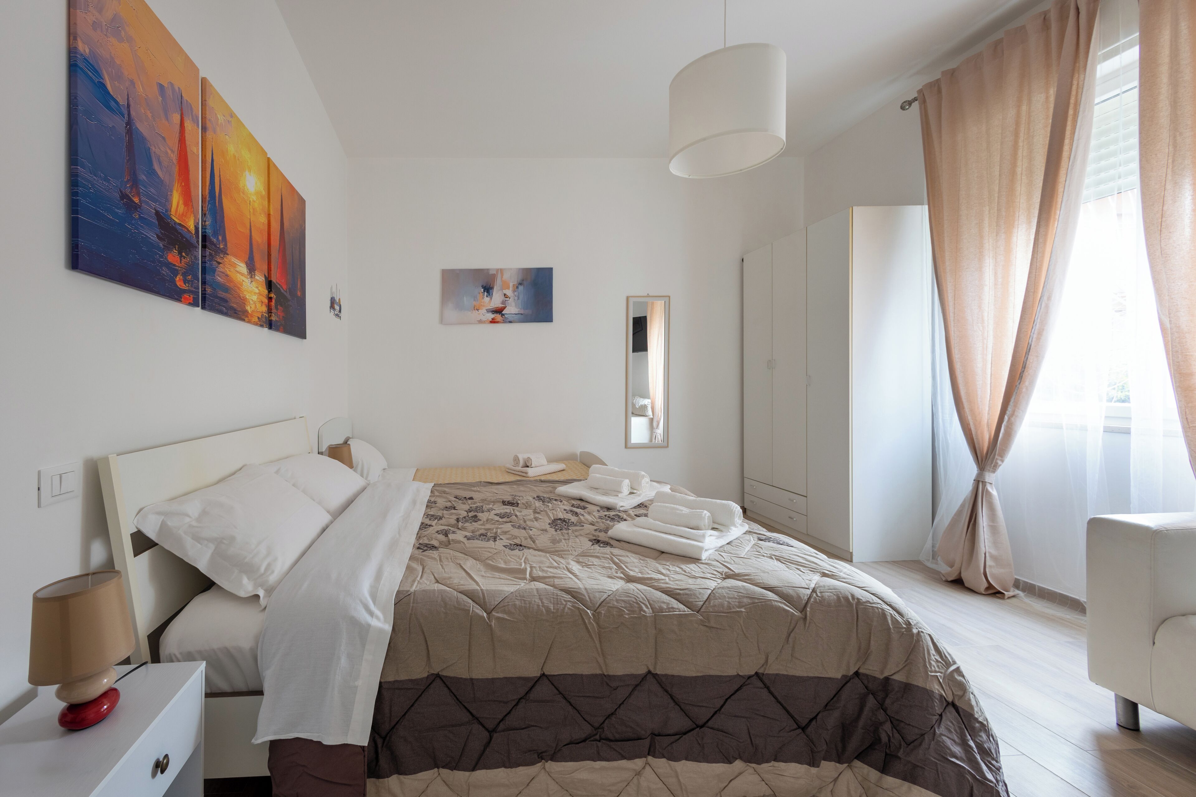 Una habitación de hotel moderna con cama, mesita de noche, lámpara, un cuadro, espejo y ventana con cortinas.