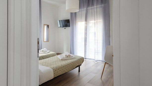 A hotel room with two beds, a flat-screen TV, a large window with curtains, and a wooden floor. - Nadia Flat - 10 minutes from the beach (Montesilvano)
