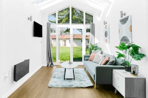 Un salon moderne avec une grande fenêtre, un téléviseur à écran plat, un canapé gris, une petite table ronde et une plante en pot.