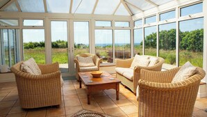 A conservatory with wicker furniture, a wooden table, and large windows offering a view of greenery. - 4 Spacious Cornish Cottages near Bude - Sleeps 25 (Bude)