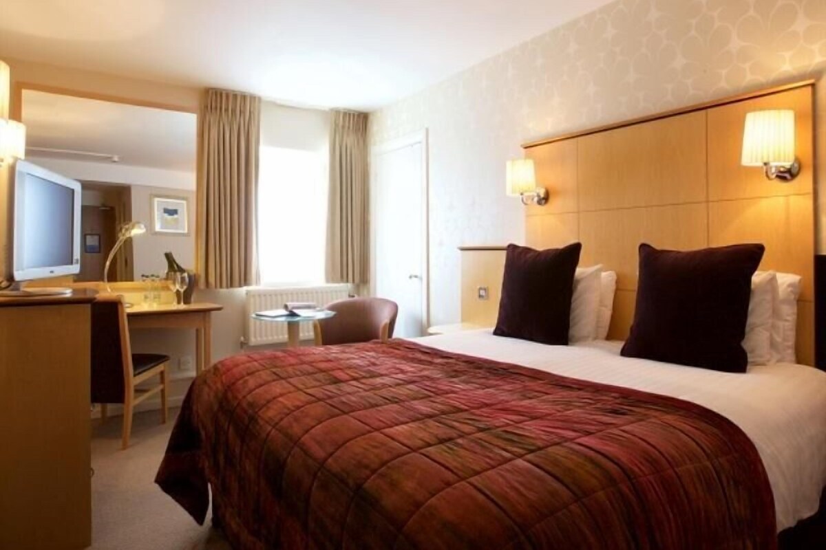 A hotel room with a bed, a desk with a computer, a chair, and a window with curtains.