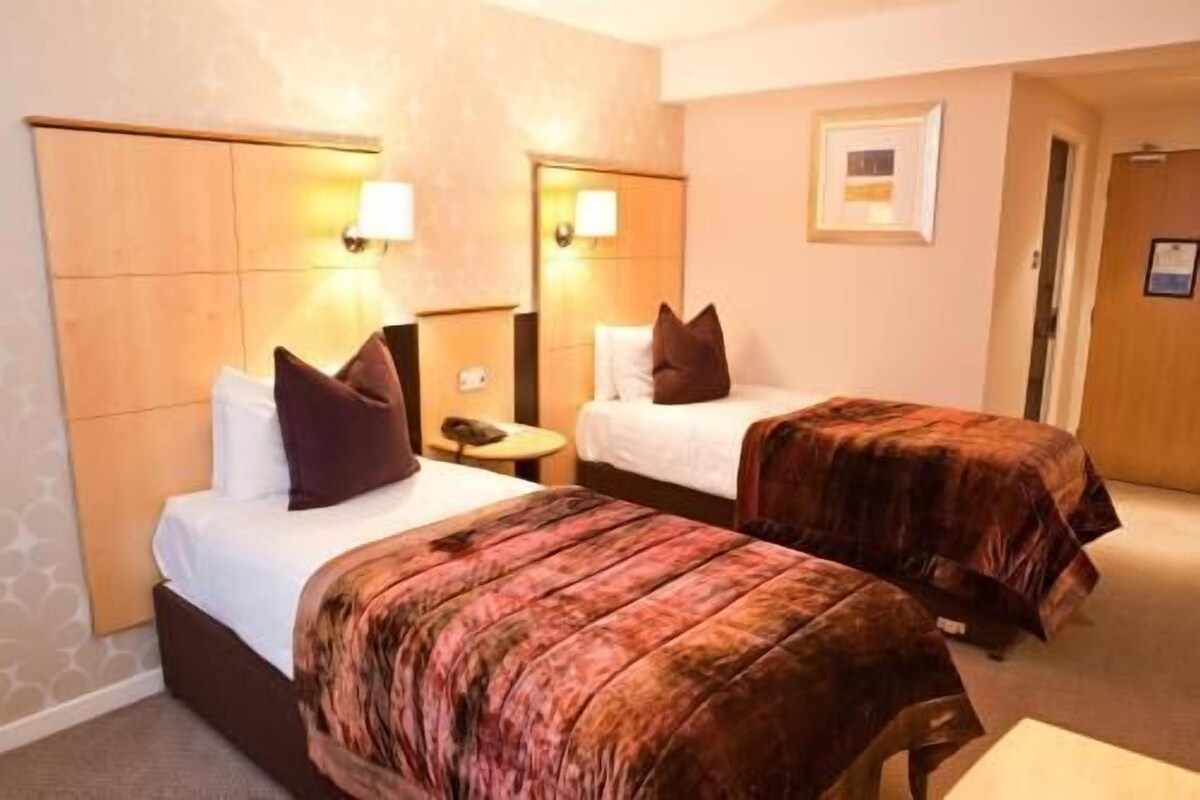 A hotel room with two beds, each with a headboard and bedside tables. There is a door leading to another room and a framed picture on the wall.