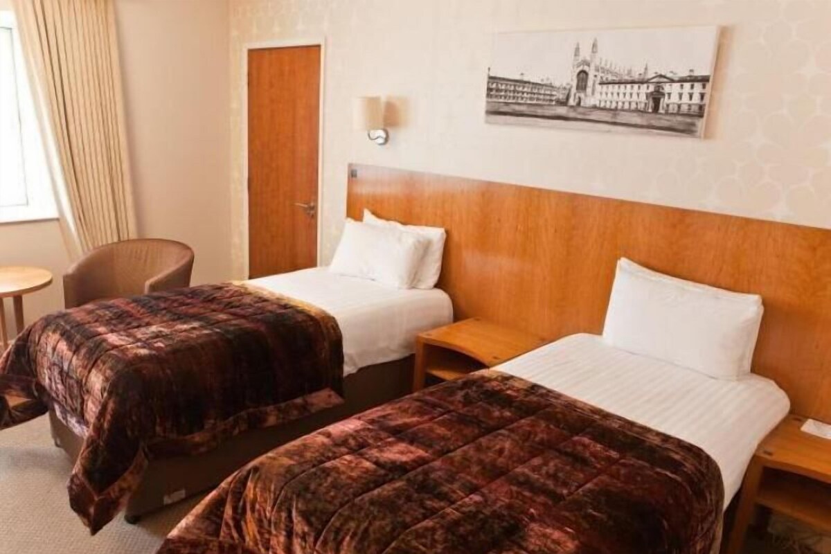A hotel room with two beds, a wooden headboard, a small table, a chair, and a painting on the wall.