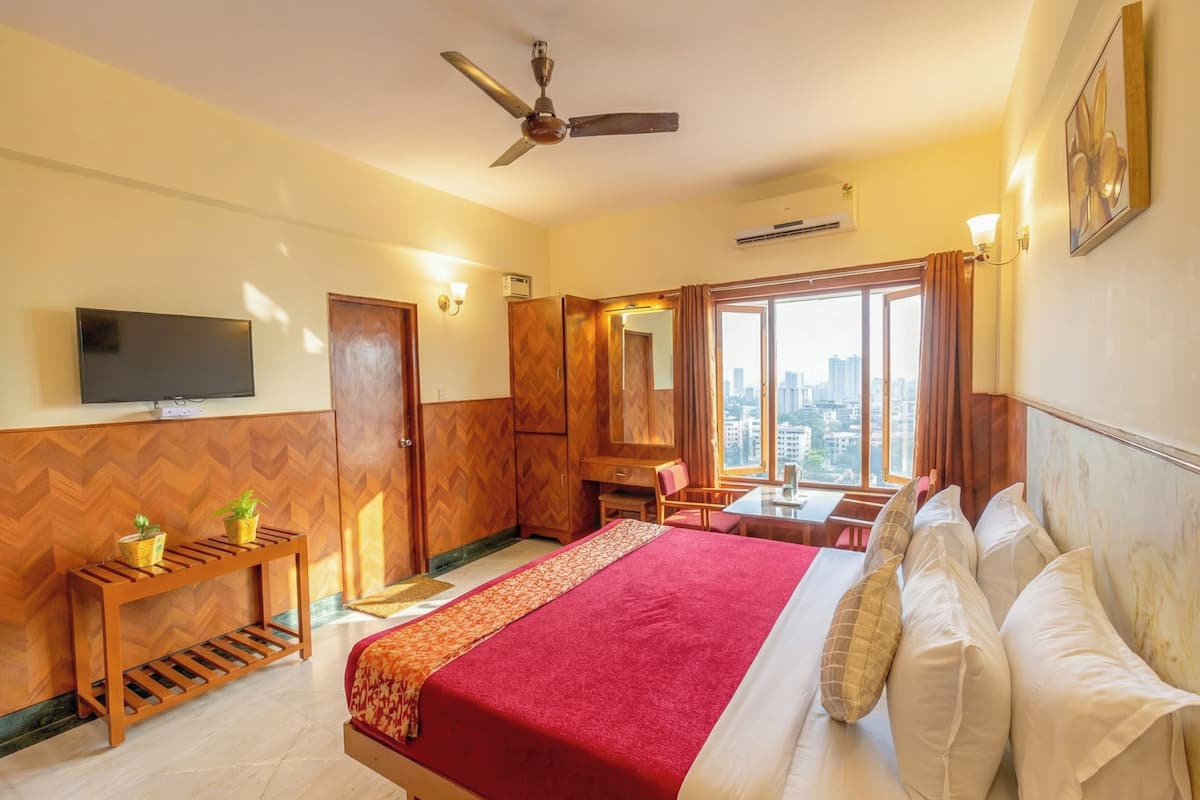 A hotel room with a bed, a TV, a desk, a chair, a ceiling fan, and a window with a city view.