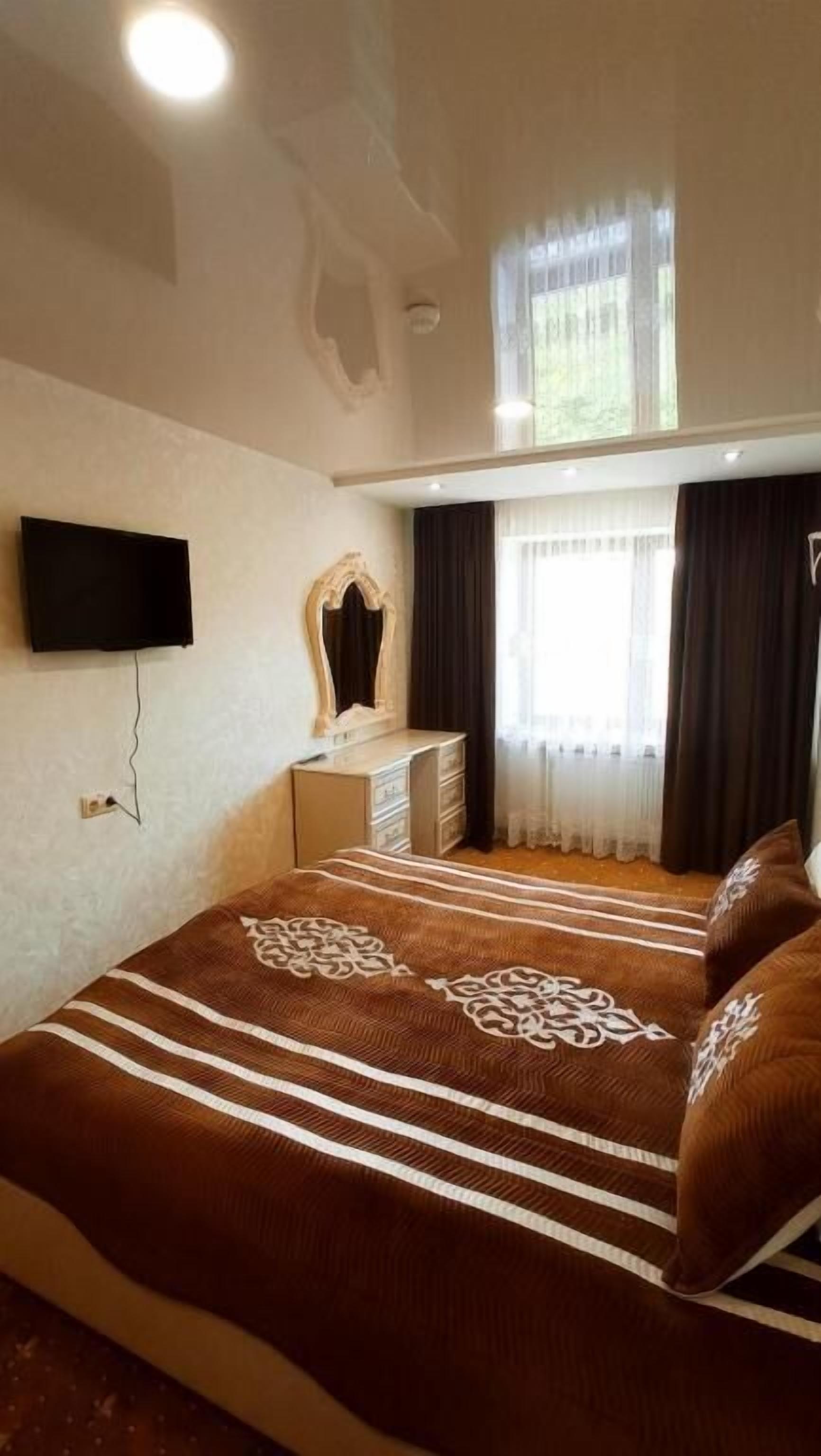 A bedroom with a bed, a dresser, a mirror, a TV mounted on the wall, and a window with curtains.