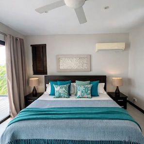 A bedroom with a bed, two bedside tables, a ceiling fan, a window with curtains, and an air conditioning unit. - Seafront 1 bedroom large penthouse apartment on the beach at Pointe Aux Biches. (Trou-Aux Biches)