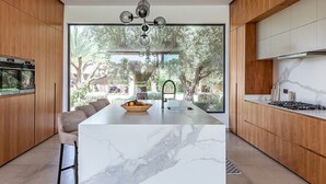 Cocina moderna con una isla de mármol, armarios de madera y un amplio ventanal con vistas a la vegetación.