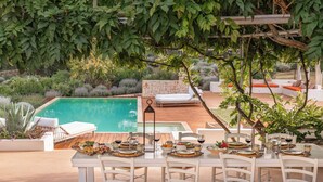 Un área de comedor al aire libre con piscina, mobiliario blanco y un muro de piedra.