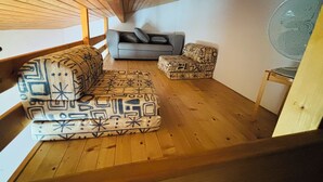 Ein Loft-Zimmer mit Holzbalkendecke, einem grauen Sofa, einem gemusterten Hocker und einem Standventilator.