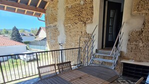 Een overdekt terras buiten, met een houten tafel en stoelen, een metalen hekwerk en uitzicht op de omgeving.
