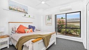 Quarto com cama, mesas de cabeceira, ventoinha no teto, uma janela grande com vista e um quadro na parede.