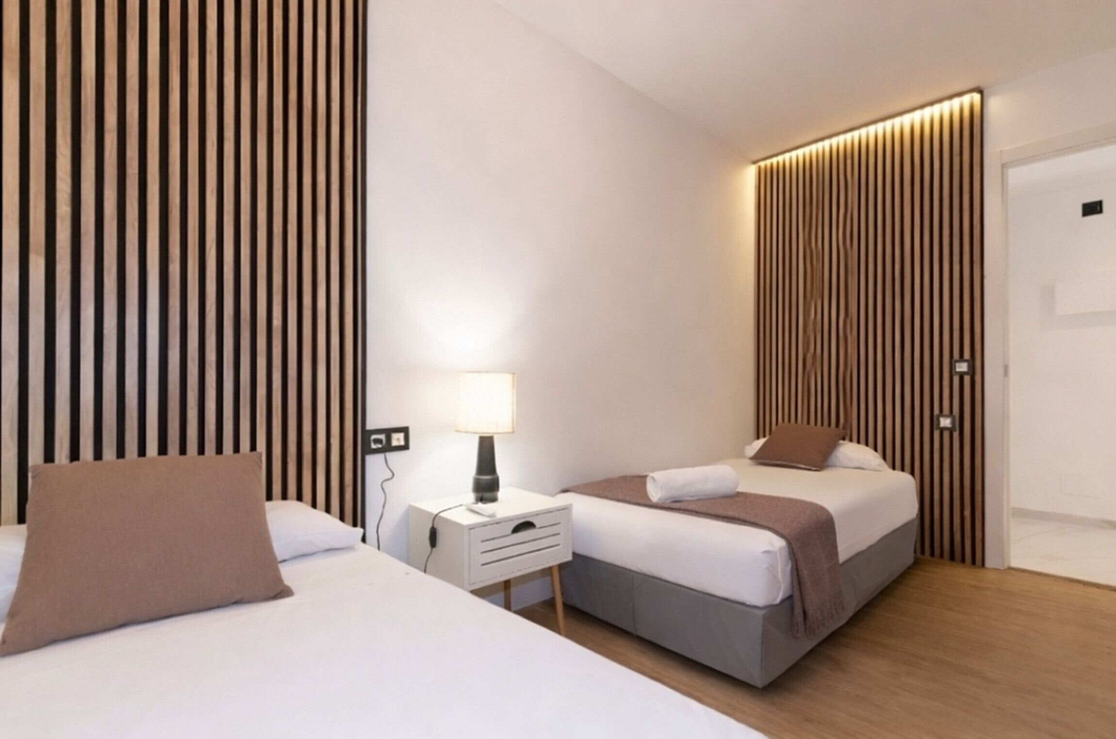 A hotel room with two beds, a bedside table with a lamp, and a wall with vertical wooden slats.