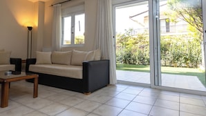 A living room with a sofa, a coffee table, and a lamp. There is a sliding glass door leading to a balcony with a view of greenery and a building.
