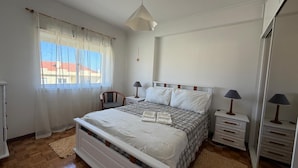 Un dormitorio con cama, mesitas de noche, una silla, armario y ventana con cortinas.