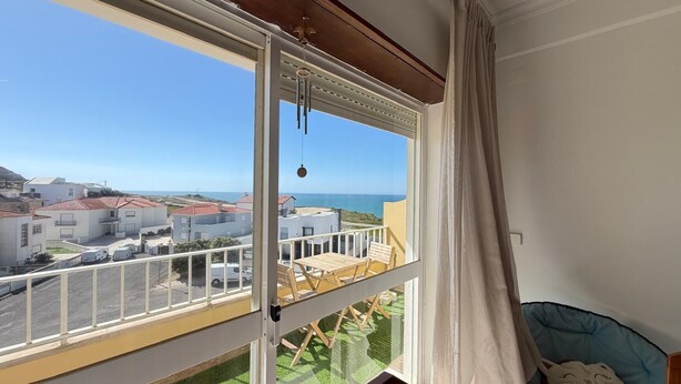 Un balcón con vista al mar y edificios, un pájaro con campanillas colgantes y una cama con cubrecolchón azul.