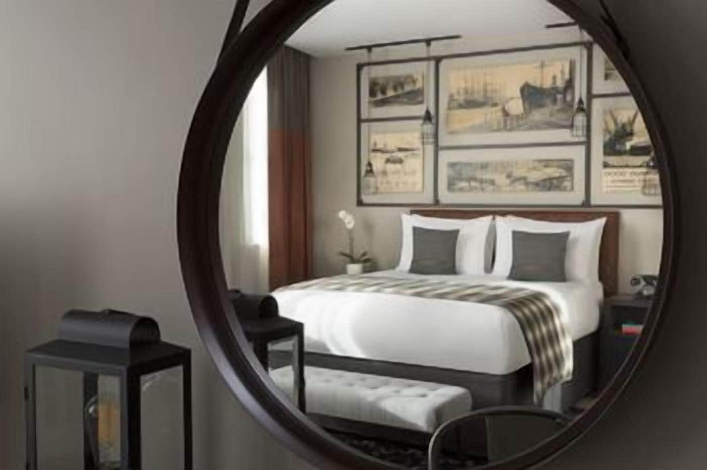 A hotel room with a bed, a bedside table, a chair, a mirror, and artwork on the wall.