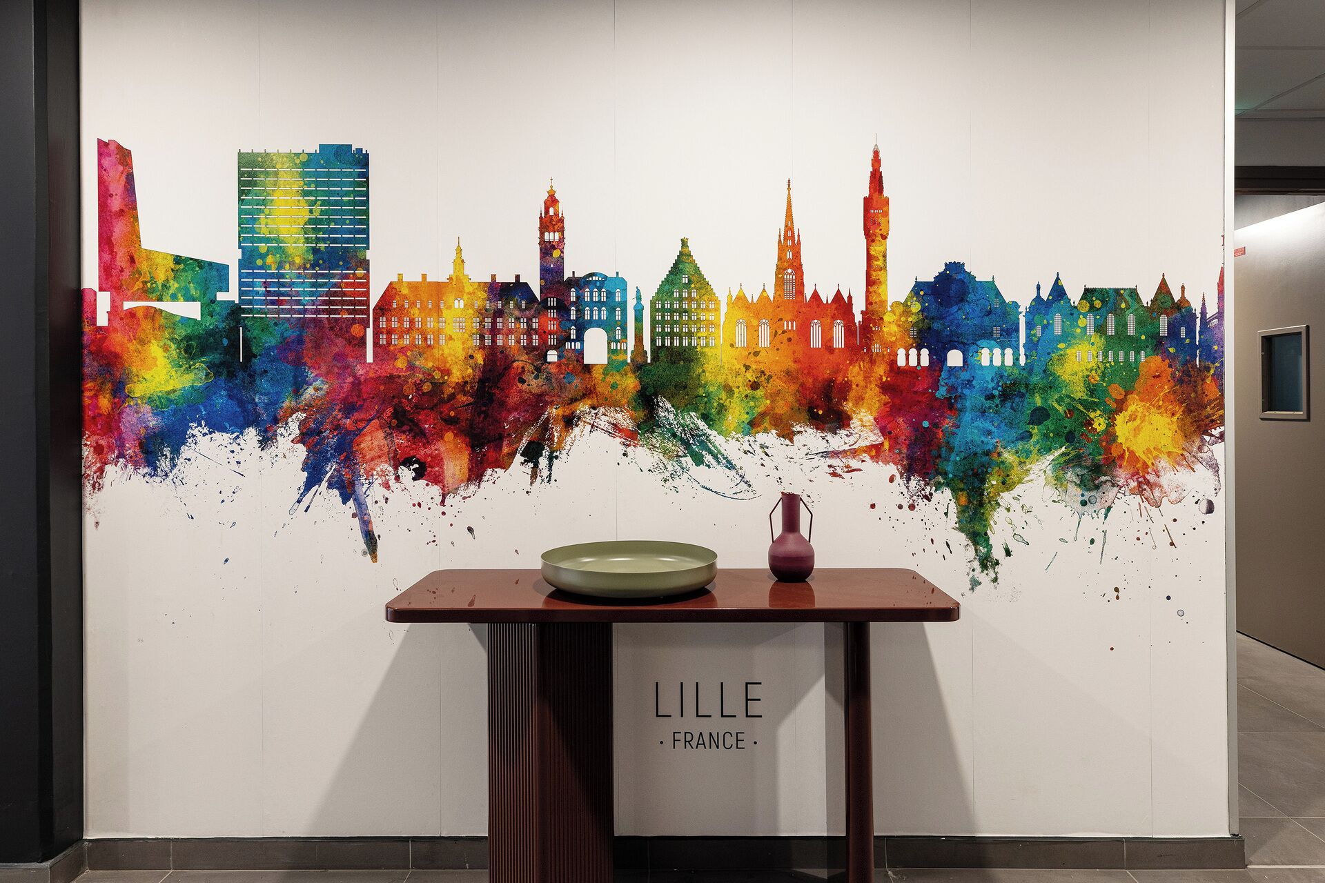 Pared con un mural colorido de un paisaje urbano, una mesa con un florero y un plato, y un letrero que dice 'LILLE FRANCE'.