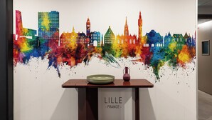 Seinässä on värikäs kaupunkimaisemaa esittävä seinämaalaus, pöydällä on maljakko ja lautanen, ja siinä on kyltti, jossa lukee: »LILLE FRANCE».
