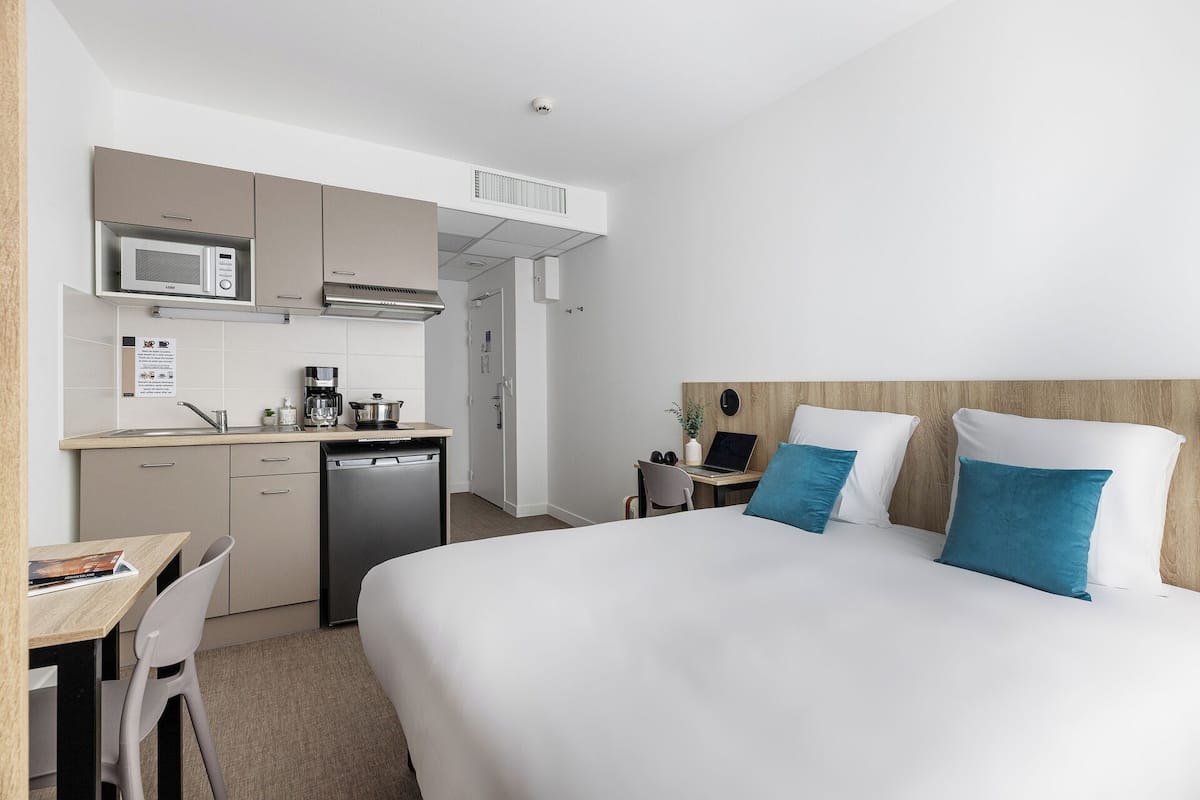 Quarto de hotel com cama, escrivaninha e cadeira, cozinha compacta com micro-ondas e cafeteira.