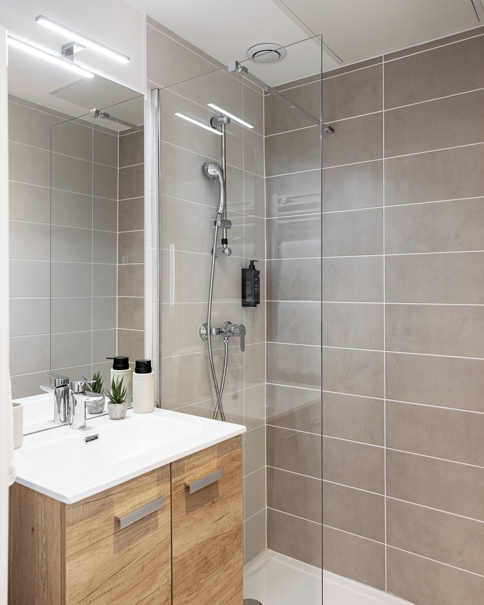 Een moderne badkamer met een douchecabine van glas, een witte wastafel met een houten kastje en een spiegel.