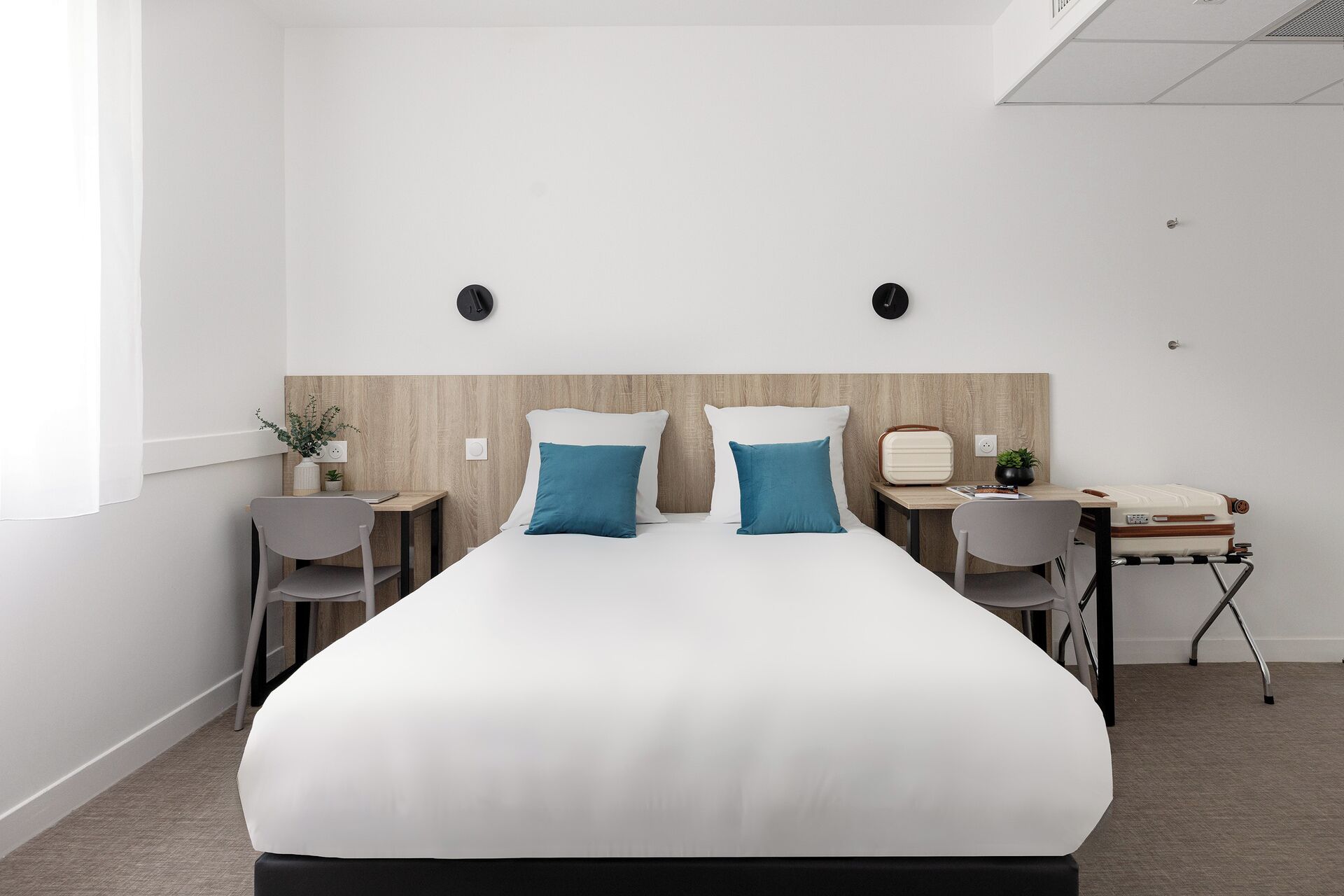 Ein modernes Hotelzimmer mit einem großen Bett, zwei Nachttischen, einem Schreibtisch mit Stuhl und einem kleinen Tisch mit Pflanze.