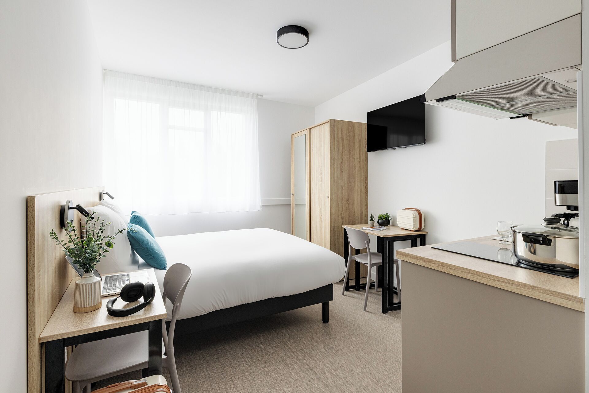 Una habitación de hotel moderna con cama, escritorio, silla, kitchenette y un gran ventanal con cortinas translúcidas.