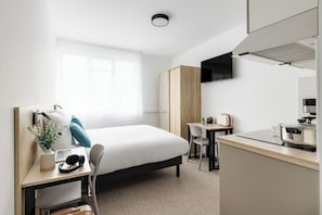 Une chambre d’hôtel moderne équipée d’un lit, d’un bureau, d’une chaise, d’une kitchenette et d’une grande fenêtre avec des rideaux légers.