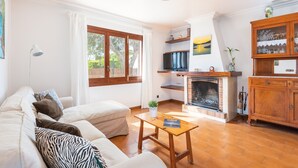 A living room with a sofa, a small table, a fireplace, and a kitchen area with a TV and cabinets. - Tolo 3 bedroom villa, Cala'n Blanes (Ciutadella de Menorca)