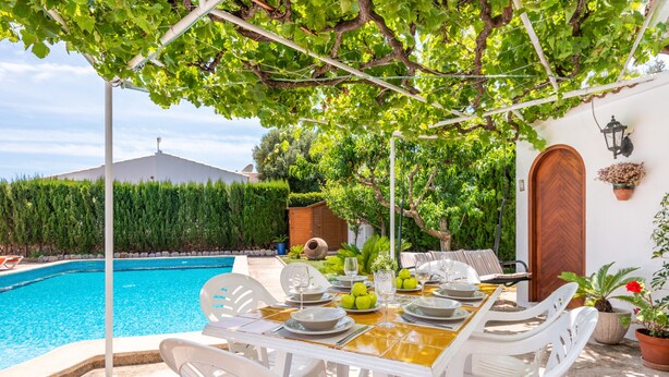 A poolside dining area with a table set for four, surrounded by white chairs, near a swimming pool. - Tolo 3 bedroom villa, Cala'n Blanes (Ciutadella de Menorca)