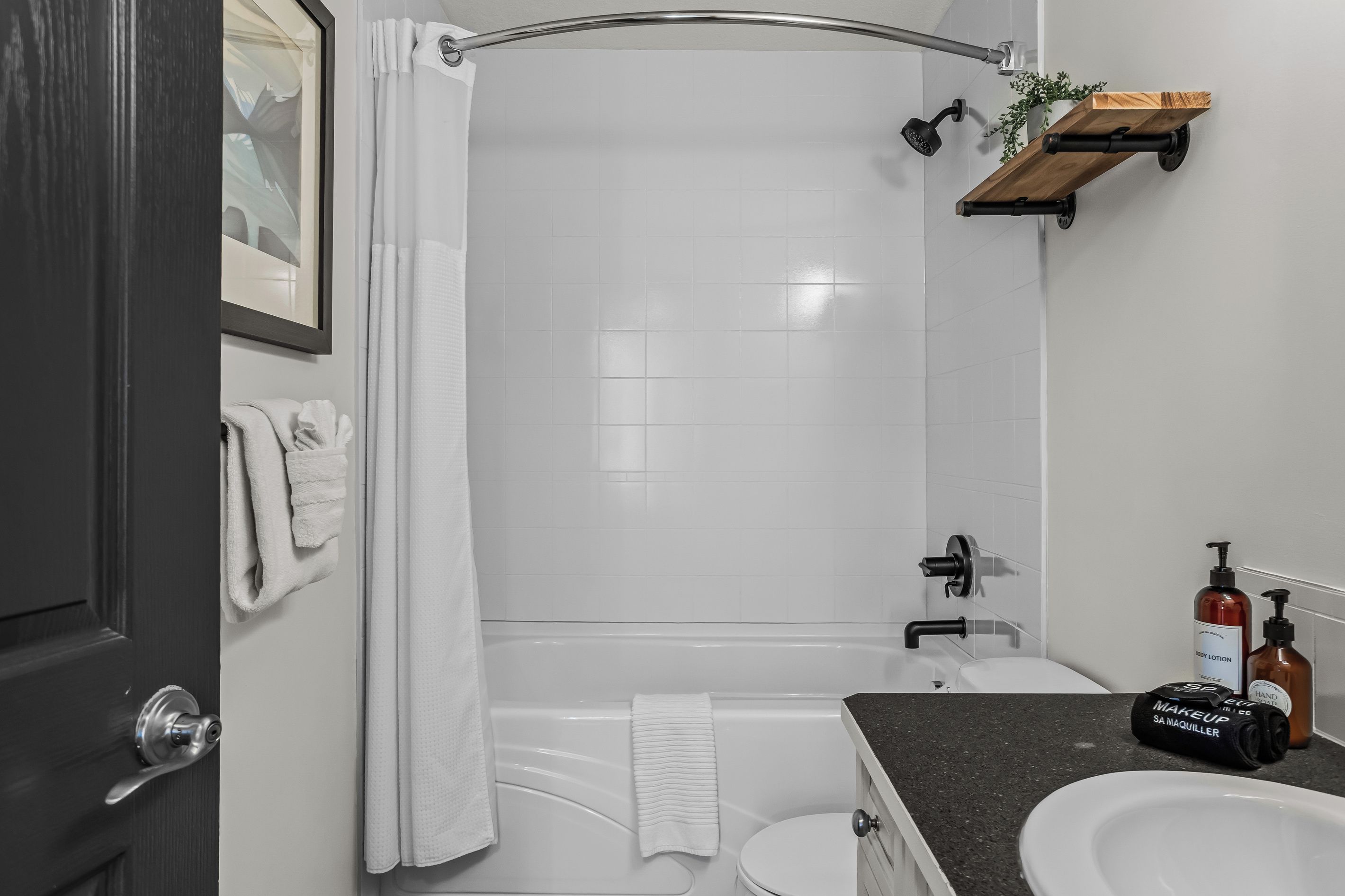 Et moderne badeværelse med badekar, bruser og toilet. Der er håndklæder hængende på væggen og en hylde med toiletartikler.