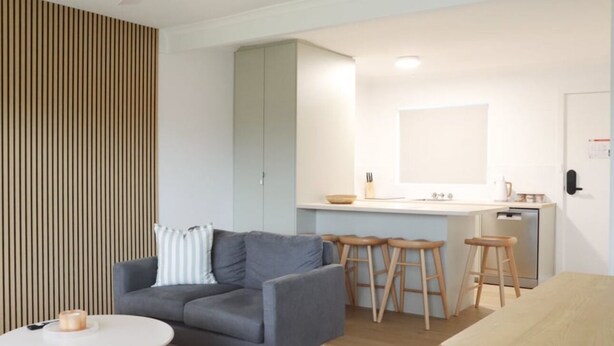 A modern living room with a grey sofa, wooden stools, a small round table, and a kitchen area with a white countertop and cabinets.