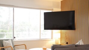 A living room with a flat-screen TV mounted on the wall, a grey sofa, a wooden chair, a small round table with a candle, and a window with a view of trees.