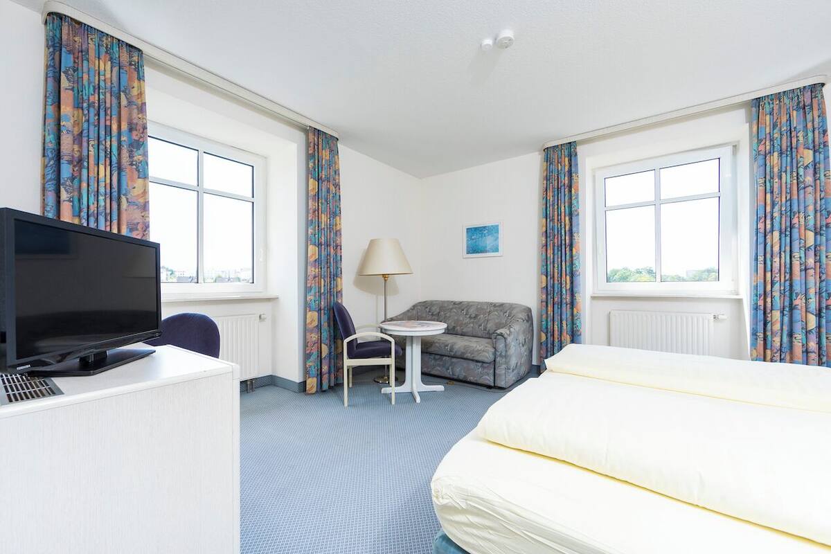 A hotel room with a bed, a desk with a television, a sofa, a chair, a small table, and curtains.