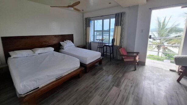 Una camera d'albergo con due letti, una ventola a soffitto, un balcone con tavolo e sedie e vista sul mare.