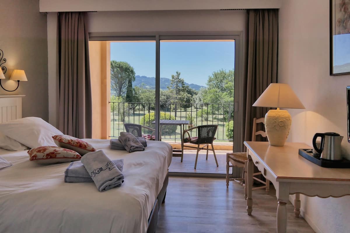 A hotel room with a bed, a desk, a chair, a table, a lamp, and a view of the outdoors.