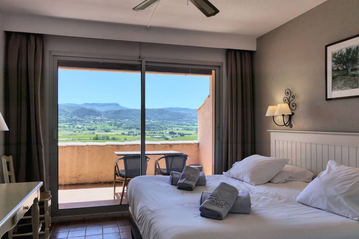 A hotel room with a bed, a desk, a chair, a balcony with a view of the countryside.