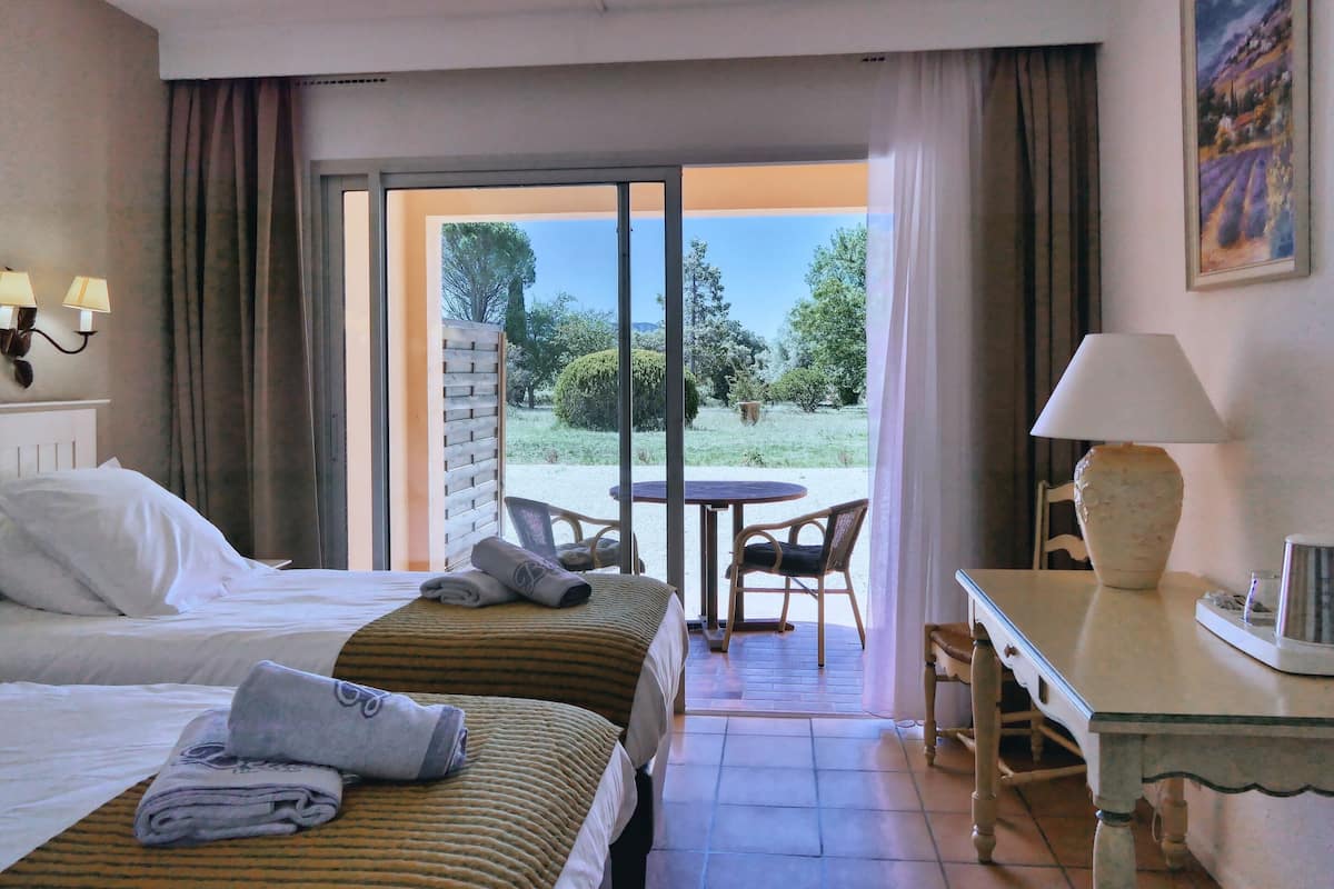 A hotel room with two beds, a dining table, chairs, a lamp, and a view of the outdoors.