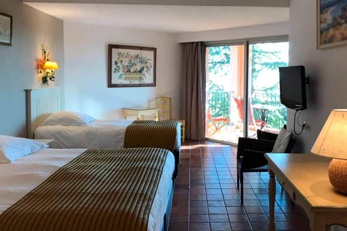 A hotel room with two beds, a desk with a lamp, a television, and a balcony with a view.