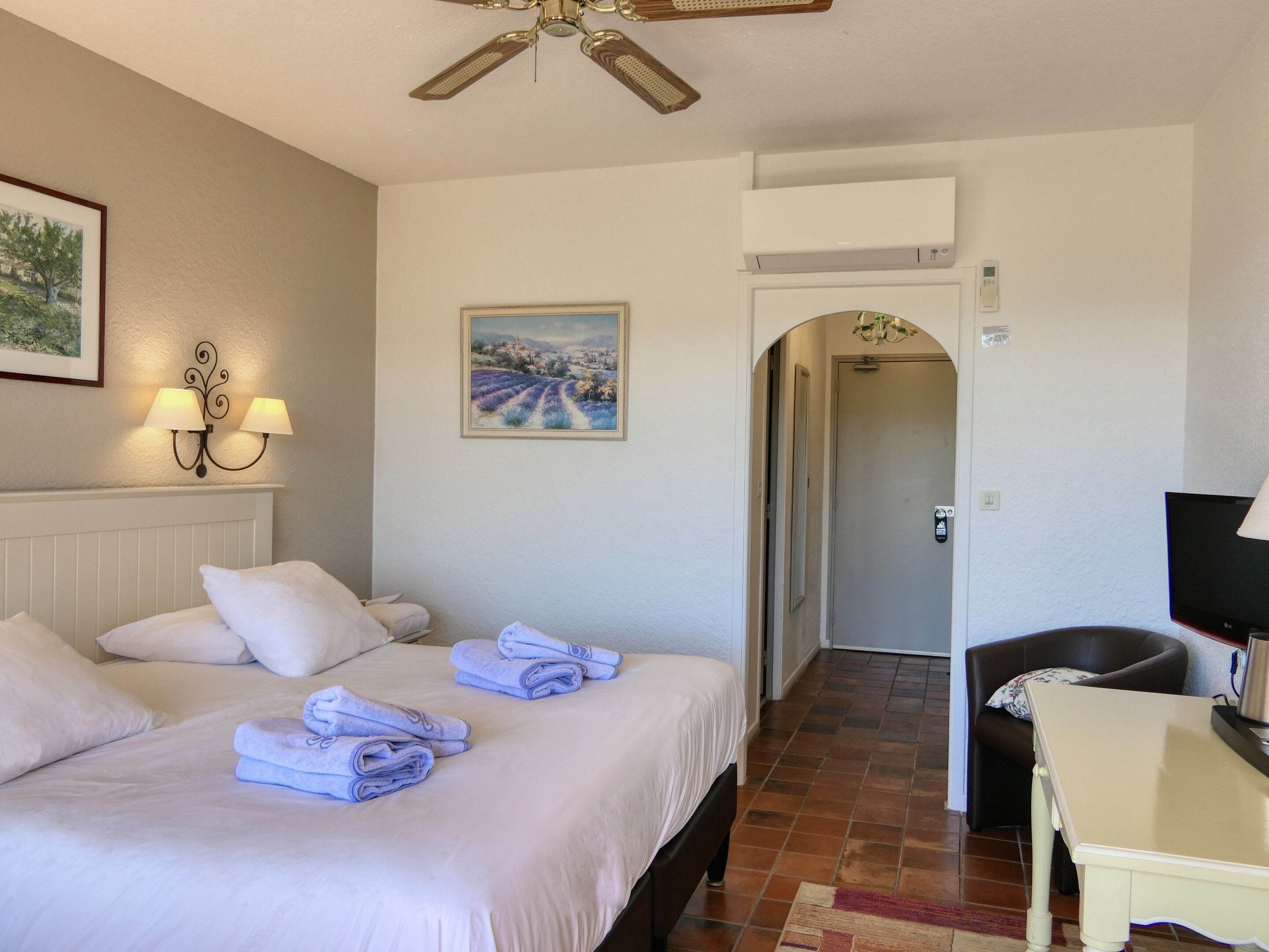 Habitación de hotel con una cama, un escritorio, una silla, un ventilador de techo, un cuadro y una puerta que da a otra habitación.