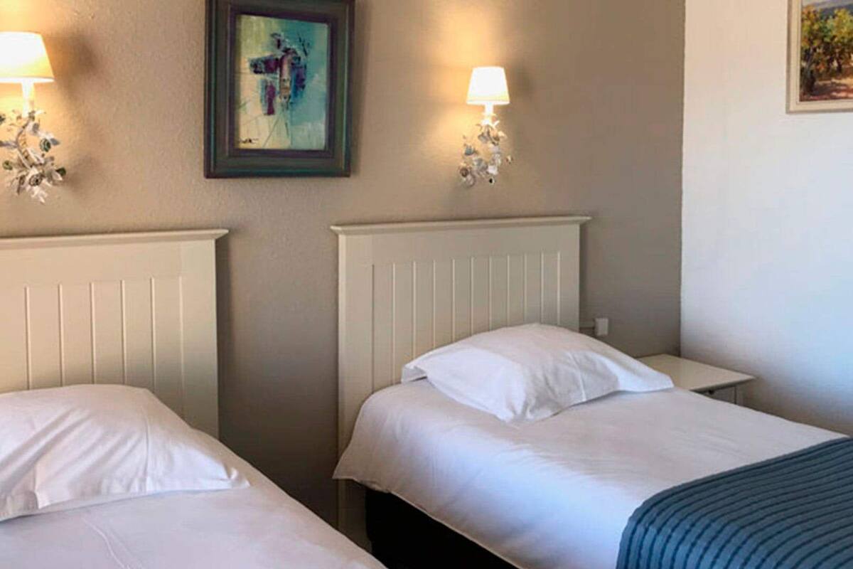Two single beds with white linens, a nightstand, a wall-mounted lamp, and a framed picture.