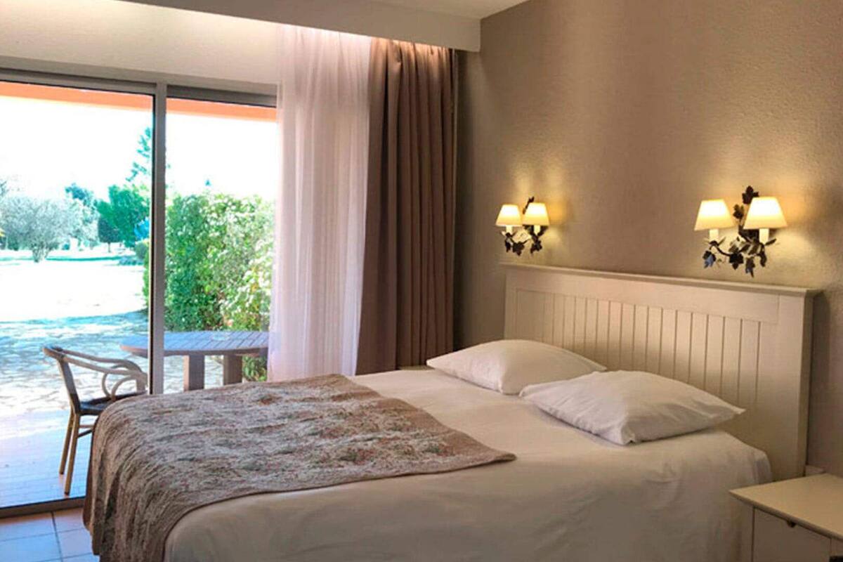 A hotel room with a bed, two wall-mounted lamps, a window with curtains, and a view of greenery outside.