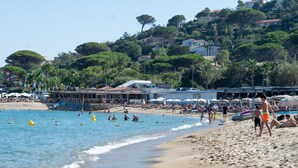 Una playa con gente nadando y tomando el sol, un edificio con un letrero 'MARIO PLAGE', y un fondo de árboles y colinas.