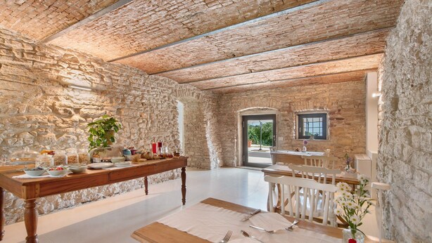 Un interior rústico con paredes de piedra, una mesa de madera dispuesta para una comida y una vista al exterior a través de un arco.
