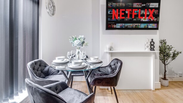 Uma área de jantar moderna com mesa de vidro, cadeiras pretas, uma planta em vaso e uma TV fixada na parede exibindo Netflix.
