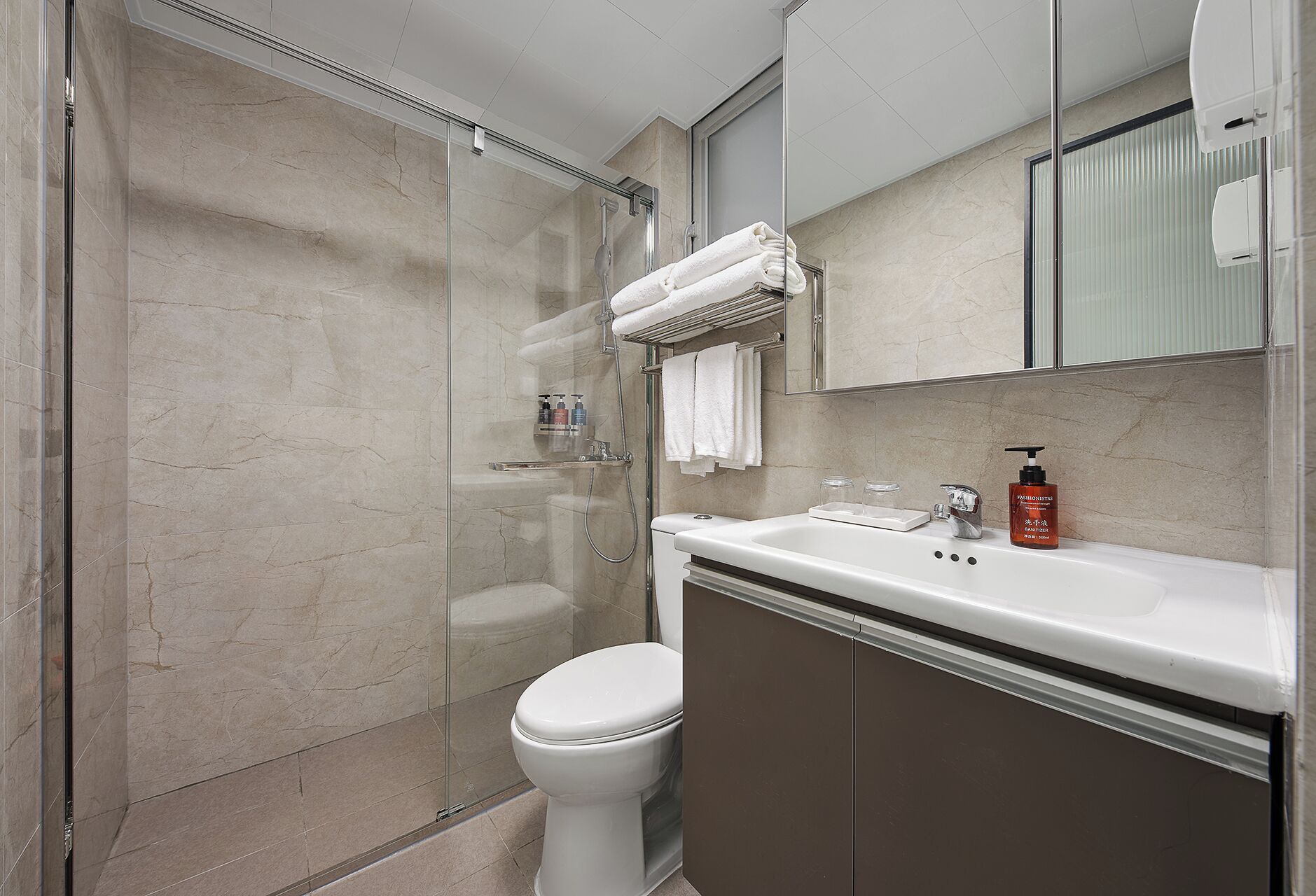A modern bathroom with a glass-enclosed shower, a white toilet, toilet, and a vanity with a mirror and a bottle of hand sanitizer.