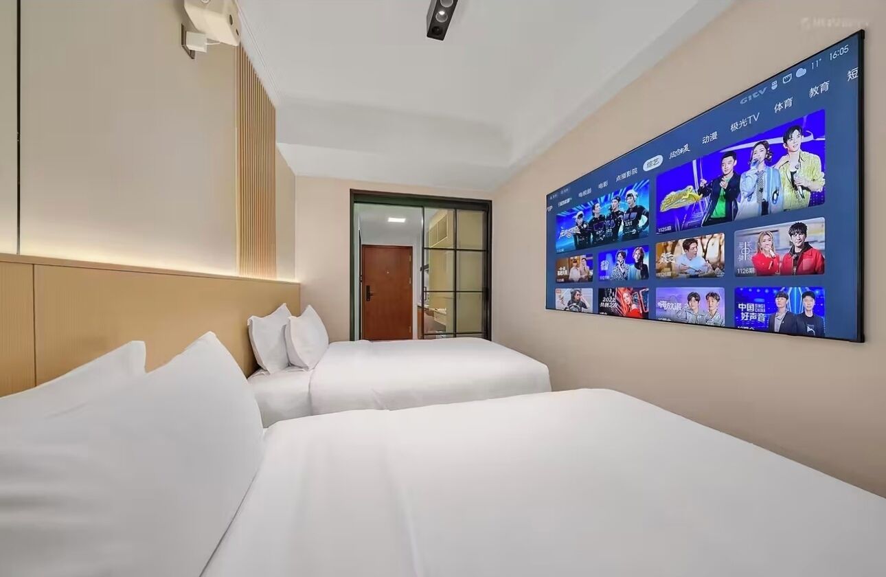 Habitación de hotel con dos camas, un televisor grande de montaje en la pared que muestra varios programas y una puerta corrediza que da a otra habitación.