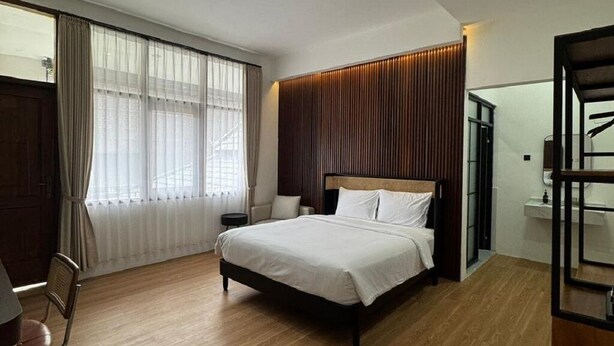 A modern bedroom with a large bed, a wooden floor, a window with sheer curtains, and a wooden headboard. - And Living Hotel (Depok)